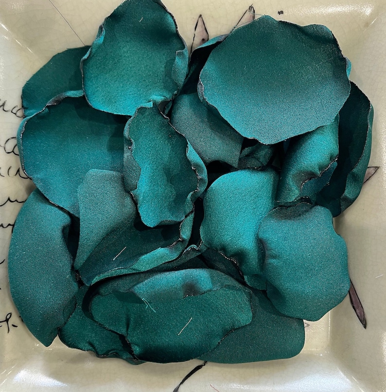 Op de afbeelding: Een verzameling tealgroene zijden bloemblaadjes, gerangschikt in een kom. De bloemblaadjes zijn zacht en delicaat, met een lichte glans.