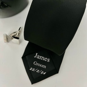 Może przedstawiać: Czarny krawat z sześciokątną etykietą z napisem "James Groom 22/5/24".