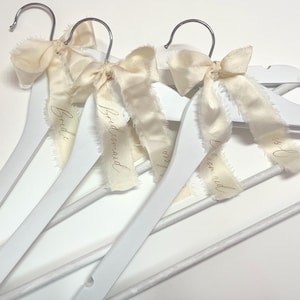 May include: A set of white wooden clothes hangers, each featuring a silver metal hook and a cream-colored frayed ribbon tied into a bow. The ribbons are printed with gold cursive text including the words Bride, Bridesmaid, and names.