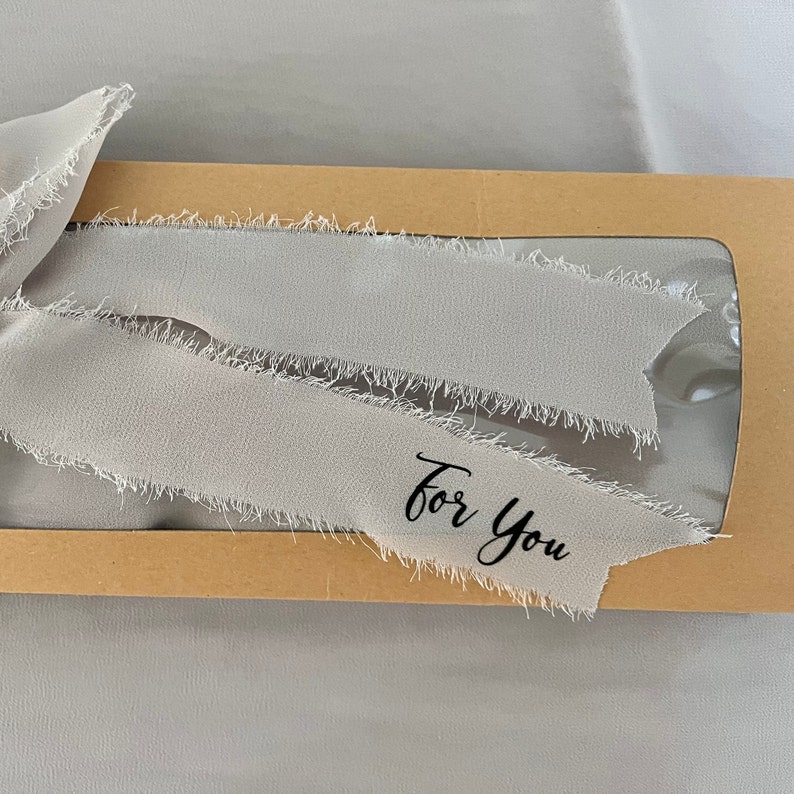 May include: A brown cardboard box with a clear window, decorated with a gray fabric ribbon with frayed edges. The ribbon has the words "For You" written on it in black.