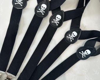 May include: Black suspenders featuring white skull and crossbones motifs. The suspenders have silver-coloured clips and are made of a robust, woven material. The skull and crossbones are on black leather-like patches.