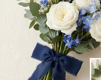 May include: A floral bouquet featuring white roses, blue delphiniums, and eucalyptus leaves, bound with a navy blue ribbon. The arrangement includes various green foliage, presenting a fresh and elegant appearance.