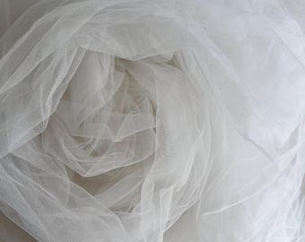 May include: A close-up of a roll of white tulle fabric. The fabric is loosely arranged, creating soft folds and shadows. The sheer material is tightly woven, and the overall effect is delicate and airy.