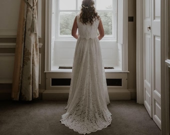 May include: A bride wearing a white lace wedding dress with a long train, standing in front of a window looking out at a view of trees.