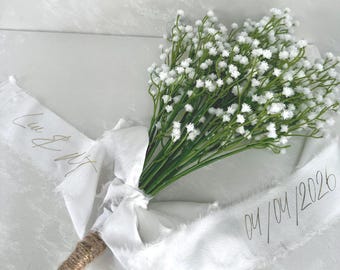 May include: A bouquet of white baby's breath flowers tied with a white frayed ribbon. The ribbon features the text "Liu & W" on one side and "04/04/2026" on the other. The stem is wrapped in brown twine, creating a rustic appearance for this floral arrangement.