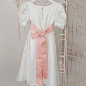 Könnte beinhalten: Ein weißes Satin-Kleid mit einem rosa Satinband, das hinten zu einer Schleife gebunden ist. Das Kleid hat einen runden Ausschnitt und kurze, bauschige Ärmel. Das Band hat ein kleines gesticktes Design.