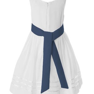 May include: A white sleeveless dress with a navy blue sash tied at the waist. The dress has a full skirt with horizontal, sheer stripes near the hem. The dress features a zip closure at the back.