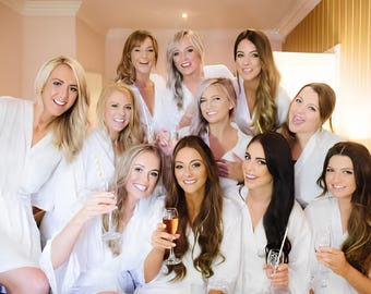 May include: A group of eleven individuals wearing matching white satin robes, each featuring a wrap-style front with a tie waist. Several individuals are holding clear glass flutes filled with a light-coloured beverage, posing together for a photo.