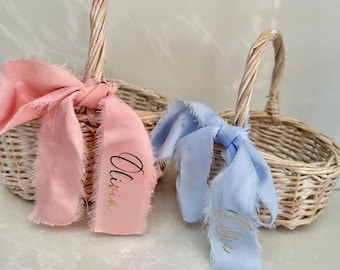 May include: Two woven wicker baskets with handles. One basket has a pink ribbon with the name "Olivia" in gold script. The other basket has a light blue ribbon with a name in gold script. Both ribbons have frayed edges.