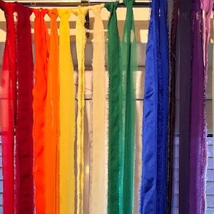 Pode incluir: Uma vibrante série de tiras de tecido em arco-íris penduradas numa barra de metal. As cores variam do vermelho ao laranja, amarelo, creme, verde, azul e roxo. As bordas do tecido são ligeiramente desfiadas.