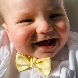 May include: A baby wearing a white shirt with a yellow bow tie smiles with a big toothy grin.