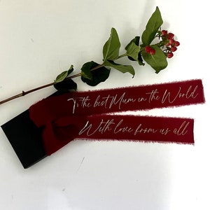 Könnte beinhalten: Zwei rote Satinbänder mit ausgefransten Kanten. Das obere Band trägt die Aufschrift "To the best Mum in the World". Das untere Band trägt die Aufschrift "With love from us all".