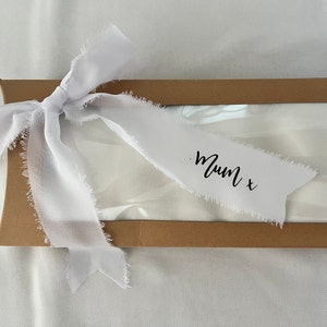 May include: A white gift box with a brown lid tied with a white ribbon. The ribbon has a frayed edge and is tied in a bow. The ribbon has the text "Mum x" written on it.