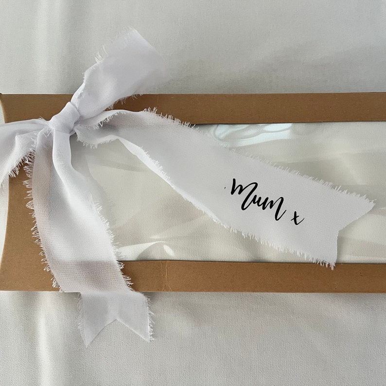 May include: A white gift box with a brown lid tied with a white ribbon. The ribbon has a frayed edge and is tied in a bow. The ribbon has the text "Mum x" written on it.