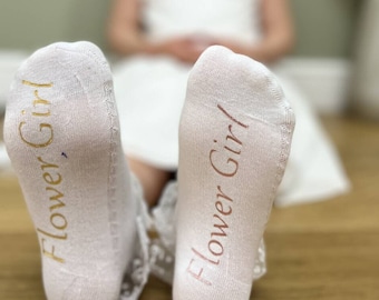 Puede incluir: Un par de calcetines blancos con ribete de encaje. Los calcetines tienen las palabras "Flower Girl" escritas en la parte inferior en letras doradas y rosas.