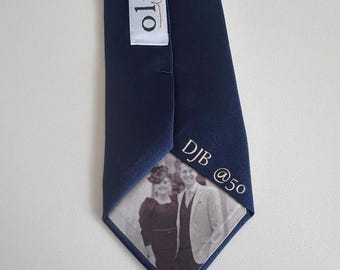 May include: A navy blue necktie featuring a white fabric label with the text olive and jane. The underside of the tie displays a black and white photograph of two people and the text DJB @50 printed in a clean, sans-serif font on the fabric.