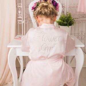 Może przedstawiać: Różowa satynowa szlafrok z napisem "Flower Girl" wysadzanym kryształkami na plecach. Szlafrok ma pasek do wiązania w talii.