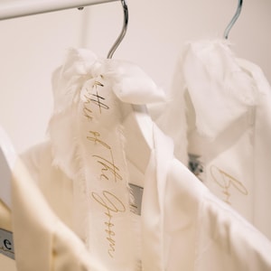 Puede incluir: Dos batas blancas colgadas en perchas con cintas de satén blancas atadas alrededor. La cinta de la bata de la izquierda tiene letras doradas que dicen "Wife of the Groom".