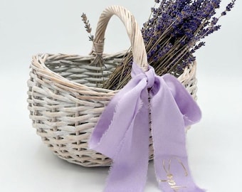 May include: A light-coloured woven wicker basket with a single arched handle, filled with dried purple lavender sprigs. A soft, light purple fabric ribbon with frayed edges is tied to the handle, featuring the name Sophie in gold cursive script.