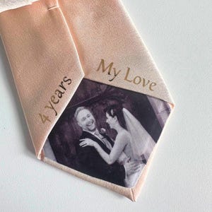 May include: A pale peach-coloured necktie featuring a custom printed black and white wedding photograph on the interior lining. The fabric is embroidered with the text My Love and 4 years in a dark brown thread on the inner flaps of the tie.