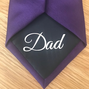 Può includere: Cravatta viola con un fazzoletto da taschino blu scuro che dice "Dad" in ricamo bianco.