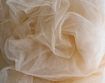 May include: Close-up of beige tulle fabric, arranged in soft folds. The fine mesh creates shadows and highlights, with a subtle sheen. The fabric appears lightweight and airy, suitable for crafting or decorative use. The colour is a warm, neutral tone.