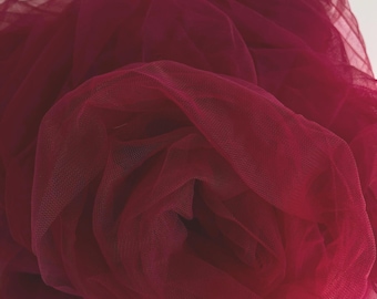 May include: Close-up of a deep red tulle fabric, bunched and layered. The fine mesh material is gathered, creating a textured, voluminous effect. The fabric is a solid, rich colour, suitable for crafting or decorative purposes.