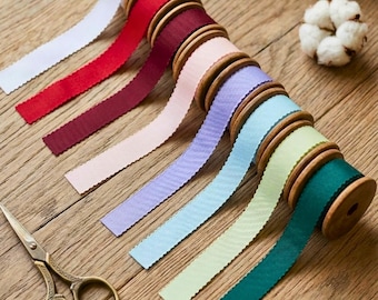 May include: A selection of colourful ribbon spools in shades of white, red, burgundy, pink, lavender, light blue, green, and teal. Each ribbon has a scalloped edge. A pair of gold scissors and a cotton ball are also present.