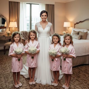 May include: A bride in a white lace and chiffon wedding dress stands with four children wearing matching pale pink satin robes and white floral crowns, each holding a small bouquet of white roses and baby's breath flowers.