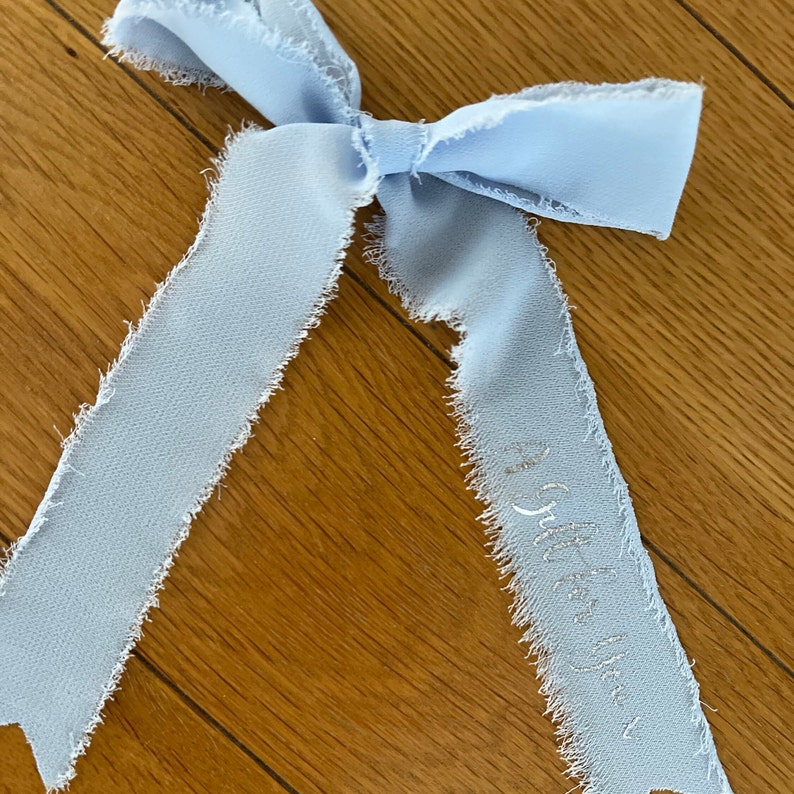 May include: A light blue ribbon with frayed edges tied into a bow. The ribbon has the words "Little One" embroidered on it in silver thread.