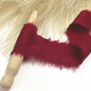 Könnte beinhalten: Ein tiefrotes Band mit ausgefransten, rohen Kanten, das um einen hellen Holzstab gewickelt ist. Das Band hat eine glatte, matte Textur und wird neben einem Büschel feiner, cremefarbener Fasern gezeigt, die hinter der Holzspule hervorragen.
