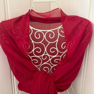 Puede incluir: Un chal de gasa rojo drapeado sobre una percha de alambre blanco. El chal es transparente y tiene una textura delicada y fluida.