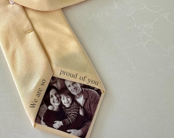 May include: A pale yellow tie featuring a family photograph. The image shows a smiling family of three. The text "We are so" and "proud of you" are printed on the tie.