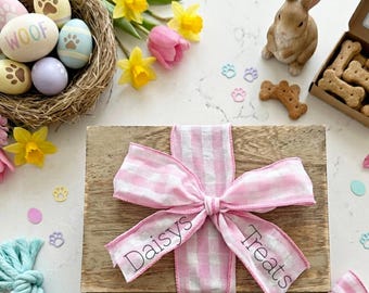 May include: A wooden box tied with a pink and white gingham ribbon, labelled "Daisys Treats". The image includes Easter eggs, flowers, a bunny ornament, and dog biscuits.