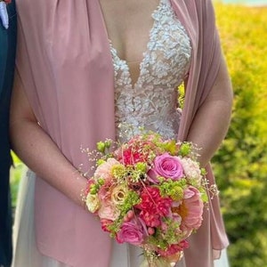 Puede incluir: Una novia sostiene un ramo de flores rosas, blancas y amarillas con follaje verde. El ramo se sostiene frente a un vestido de novia blanco con un corpiño de encaje y una chalina de gasa rosa.