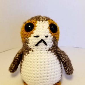May include: A crocheted stuffed toy resembling a character from Star Wars. The toy is brown, white, and orange with black eyes.