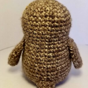 May include: A brown crocheted owl plush toy with yellow eyes. The owl is standing on a white surface.