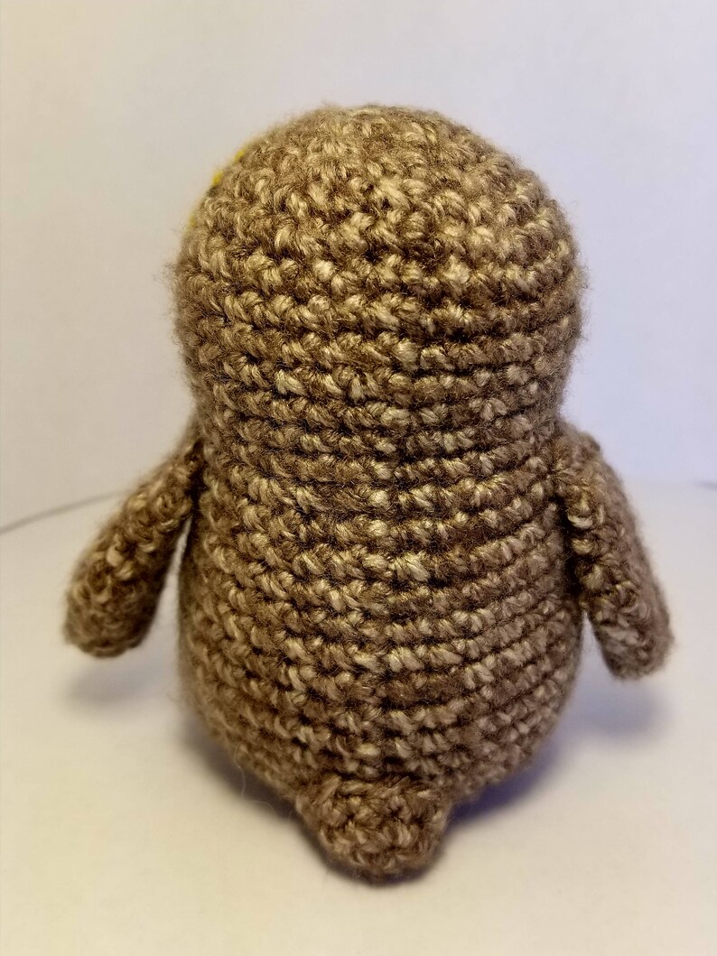 May include: A brown crocheted owl plush toy with yellow eyes. The owl is standing on a white surface.