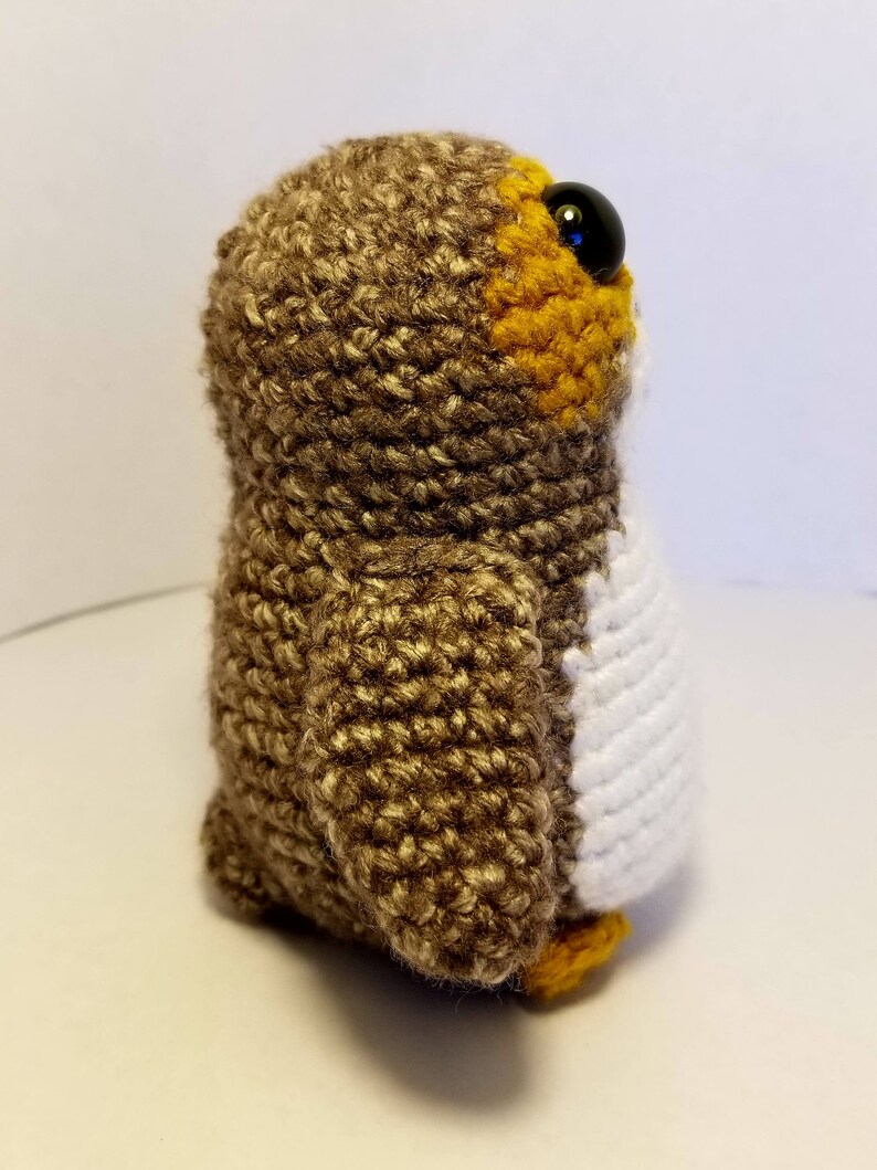 May include: A brown and white crocheted owl plush toy with black eyes. The owl has a yellow beak and feet.