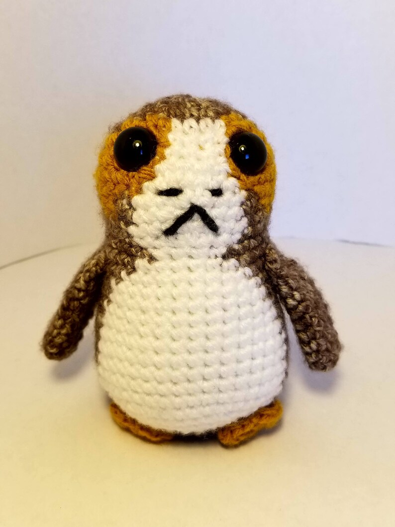 May include: A crocheted stuffed toy resembling a character from Star Wars. The toy is brown, white, and orange with black eyes.