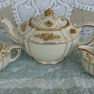 May include: A white teapot, creamer, and sugar bowl with gold trim and floral designs. The set is on a light blue tablecloth with a white lace overlay.