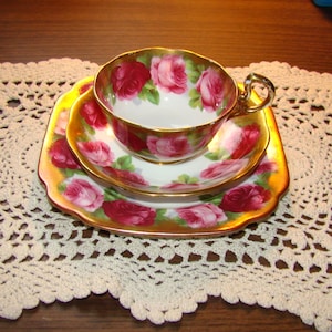 May include: A vintage tea set comprising a teacup, saucer, and plate. The set is decorated with a rose pattern in red and pink, with gold edging. The teacup has a gold handle. The set rests on a cream-coloured, crocheted doily.
