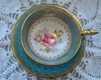 May include: A vintage teacup and saucer set with a floral design. The saucer is turquoise blue with gold accents and the teacup features a pink rose and green leaves on a white background.