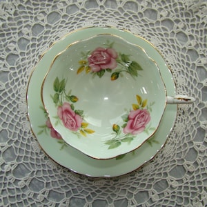 May include: A delicate teacup and saucer set with a light green background and a floral design featuring pink roses. The set has a gold trim around the edges.