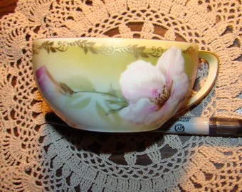 May include: A vintage teacup with a floral design. The cup features a large pink and white flower with green leaves against a yellow background. Gold trim accents the rim and handle. The cup is placed on a beige doily.