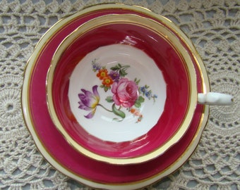 May include: A pink teacup and saucer set with a gold rim and floral design. The cup and saucer are decorated with a white background and a floral pattern in shades of pink, red, and yellow.