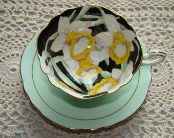 May include: A vintage teacup and saucer set with a delicate floral design. The cup features a black background with white and yellow daffodils. The saucer is a pale green colour with a gold trim.