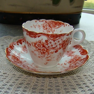 May include: A vintage teacup and saucer set with a red and white floral pattern. The cup and saucer have a gold trim and a scalloped edge.
