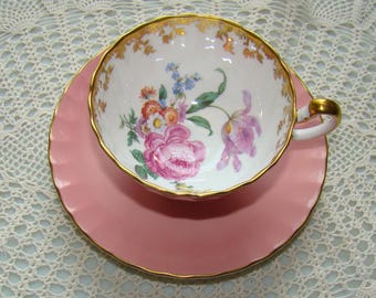 May include: A pink teacup and saucer set with a floral design. The teacup has a gold rim and the saucer has a scalloped edge.