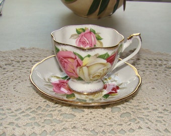 May include: A white teacup and saucer with a gold rim and floral design. The design features pink, white, and red roses with green leaves.